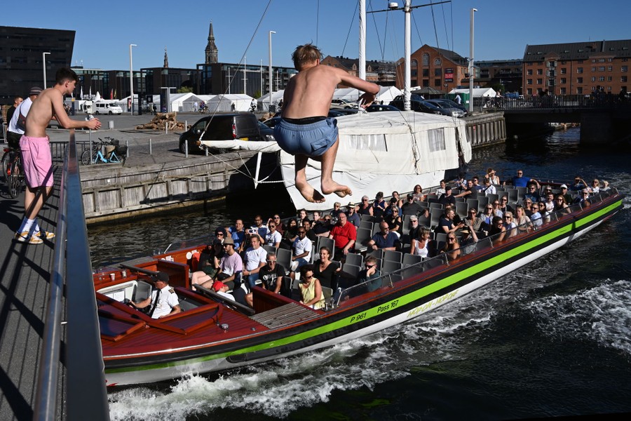 A man in a bathing suit captured midair, jumping from a bridge into a canal, as a boat full of tourists passes nearby
