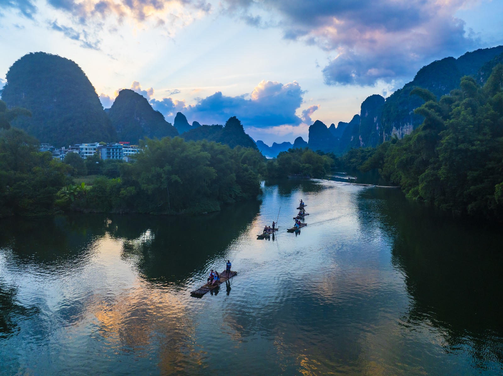 Tourists ride on several bamboo rafts down a calm river, past strikingly tall hills and rock formations.