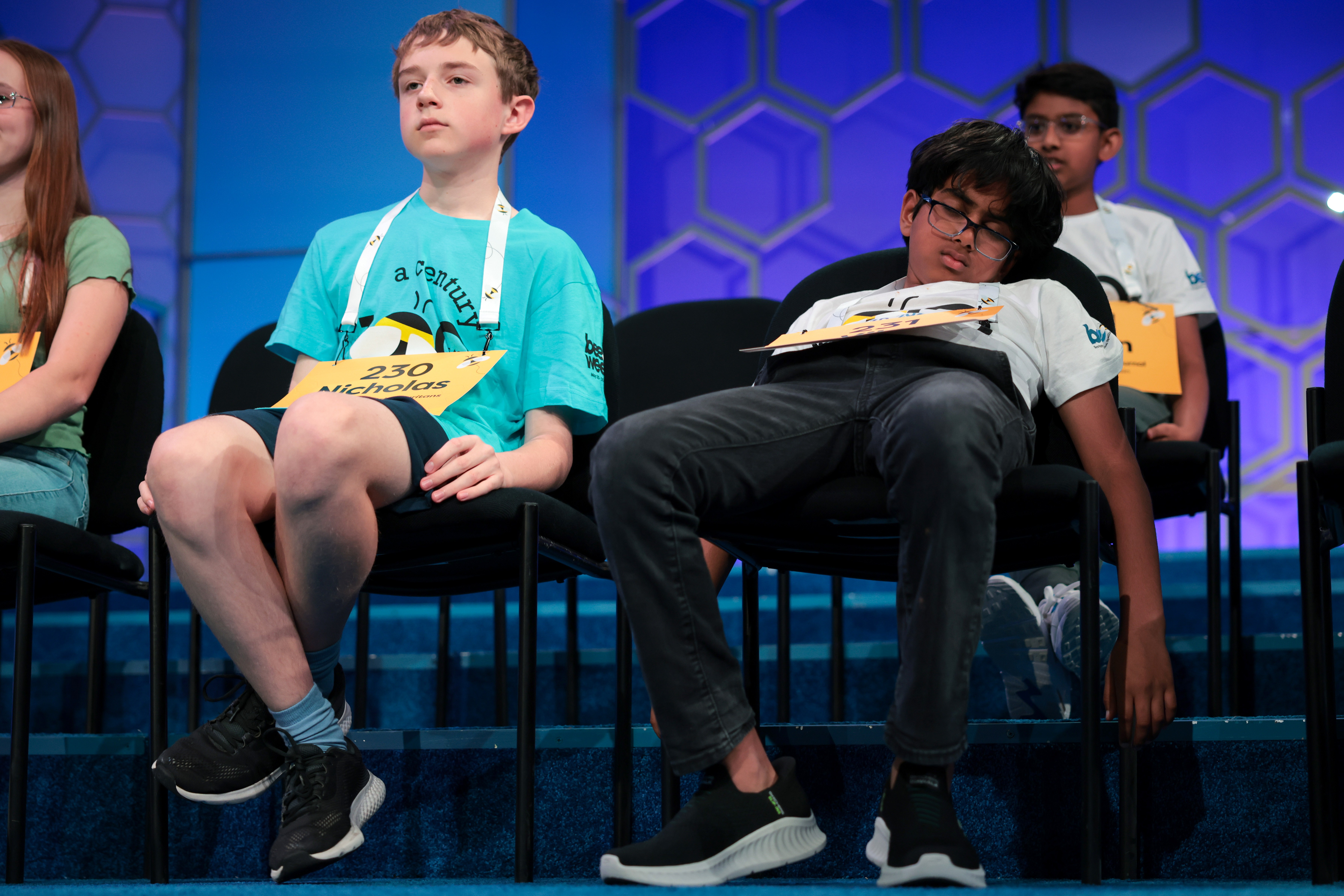Several young spelling bee contestants sat on a chair on the stage, one of whom seemed to be asleep.