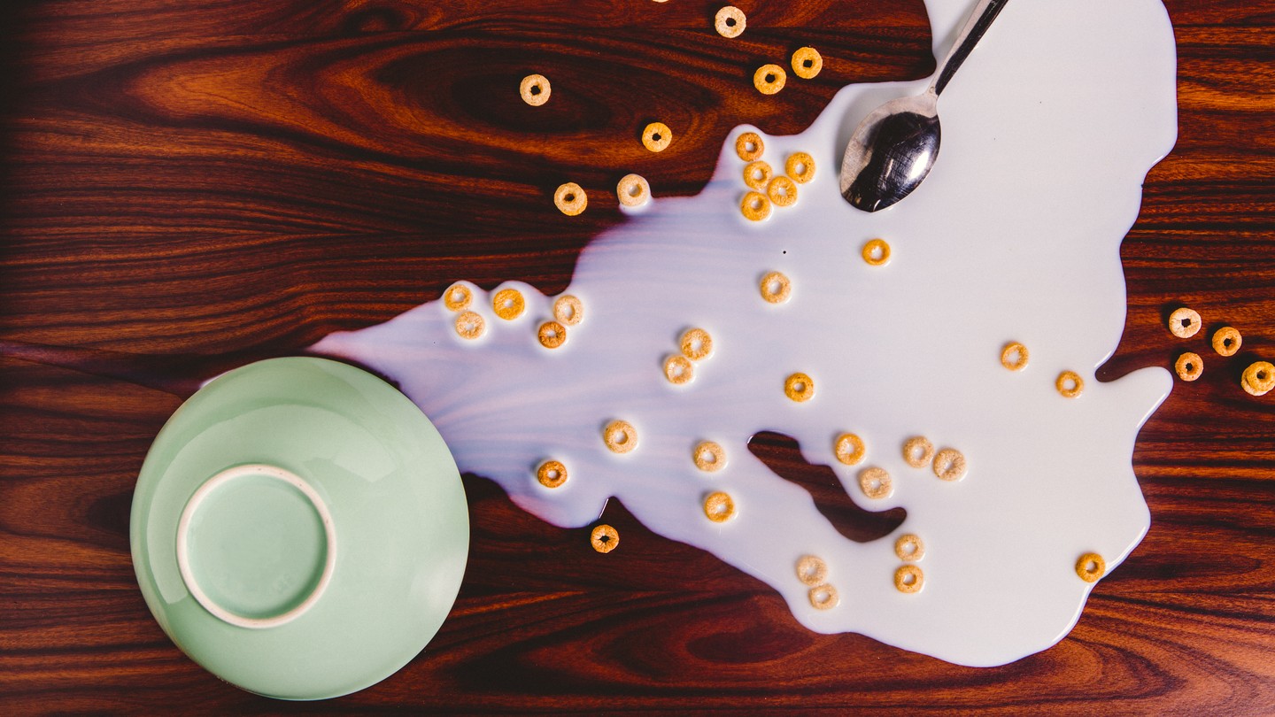 A green bowl upside down on a reddish-brown wood table, with spilled milk and Cheerios to its right. A silver spoon lies upside down in the milk.