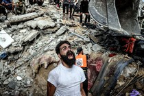 A man reacts, standing beside a huge pile of rubble following an air strike. Workers and onlookers stand nearby and atop the rubble.