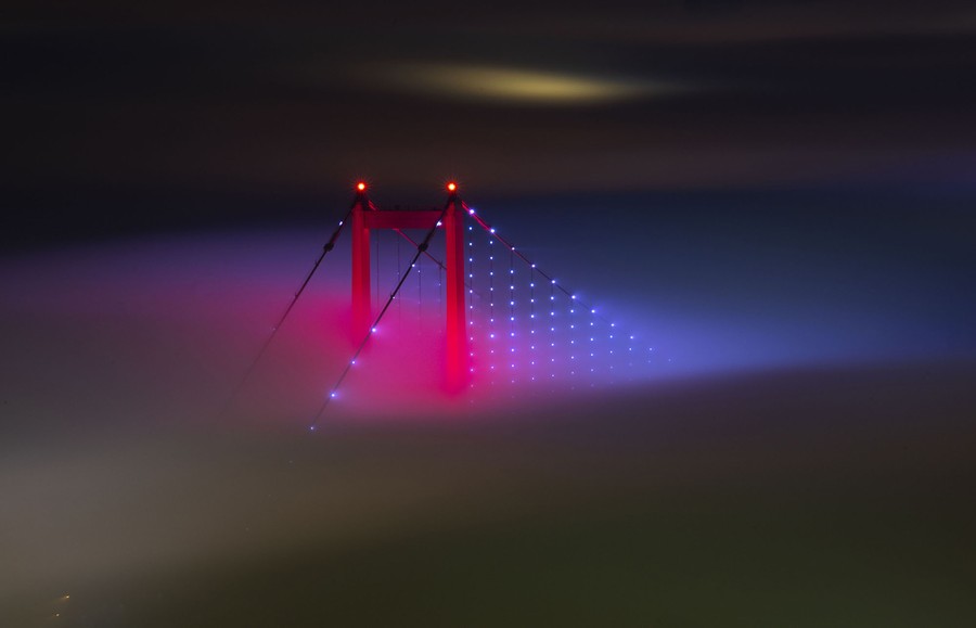 The upper portion of a bridge support pokes through a fog bank at night.