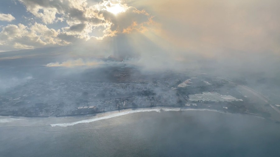 An aerial view of smoke rising from burned areas of a coastal town.