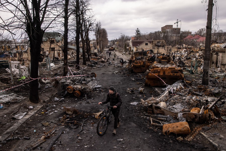 A man pushes a bike through debris and destroyed tanks on a street.