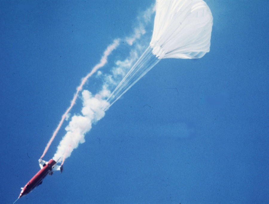 A small rocket hangs beneath a parachute, emitting a trail of smoke.