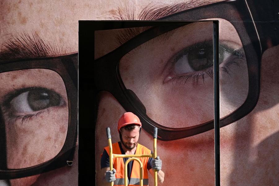 A worker stands outside a shop decorated by a large mural of a person's face with glasses.