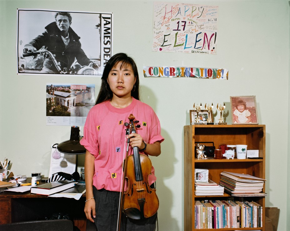 photo of girl with dark hair wearing pink shirt and holding violin and bow in left hand, standing in front of desk and bookcase with posters on wall including James Dean