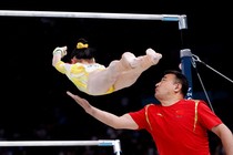 A gymnast falls face-down from a high bar she had been swinging from as a coach reaches out one arm to slow her.