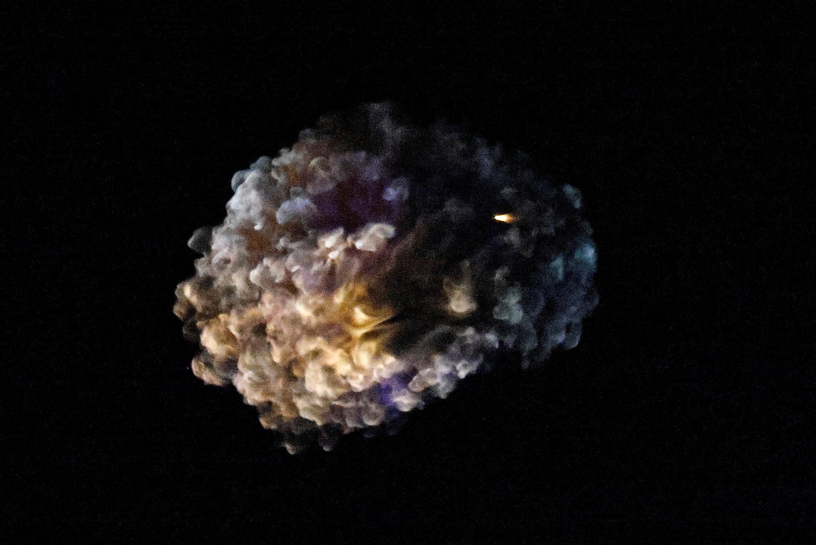 A multicolored cloud, left by an explosion, hangs in the night sky.
