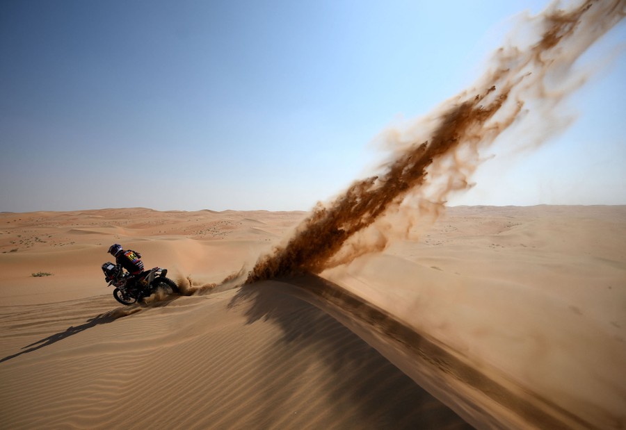 A motorcyclist kicks up a plume of sand while riding over a dune in a desert.