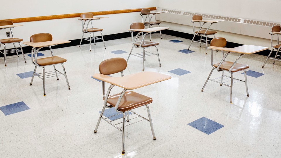 A photo of single-seat desks spaced out in a classroom with white walls and a blue-and-white-tiled floor