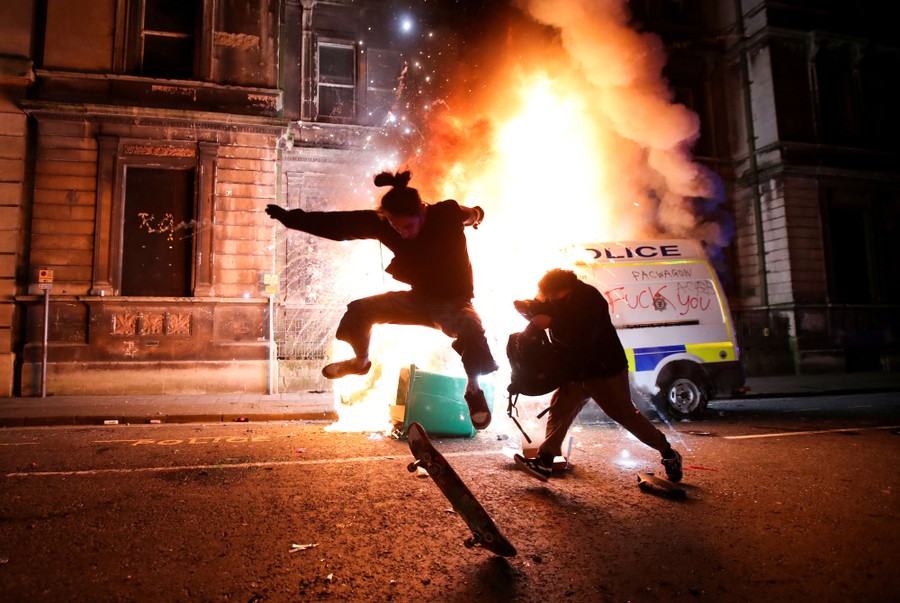 A person does a skateboard trick in front of a burning police vehicle.