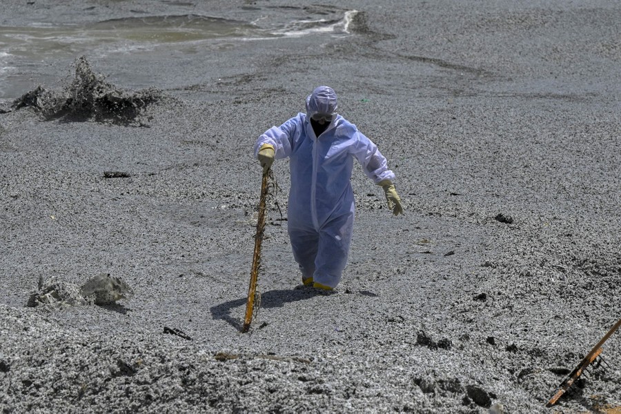 A Navy member in protective gear wades through seawater and plastic debris.