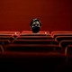 Person sitting alone in a red theater wearing a black mask