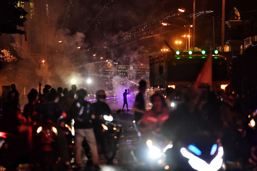Protesters and police move about in a street at night, with fireworks exploding nearby.