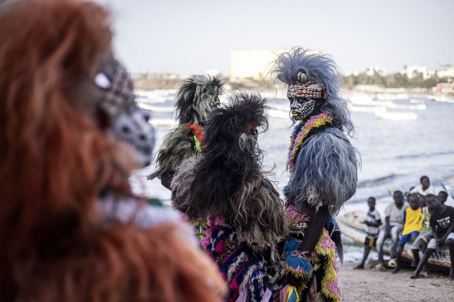 Performers wear traditional costumes of fur and intricate face paint while on a beach.
