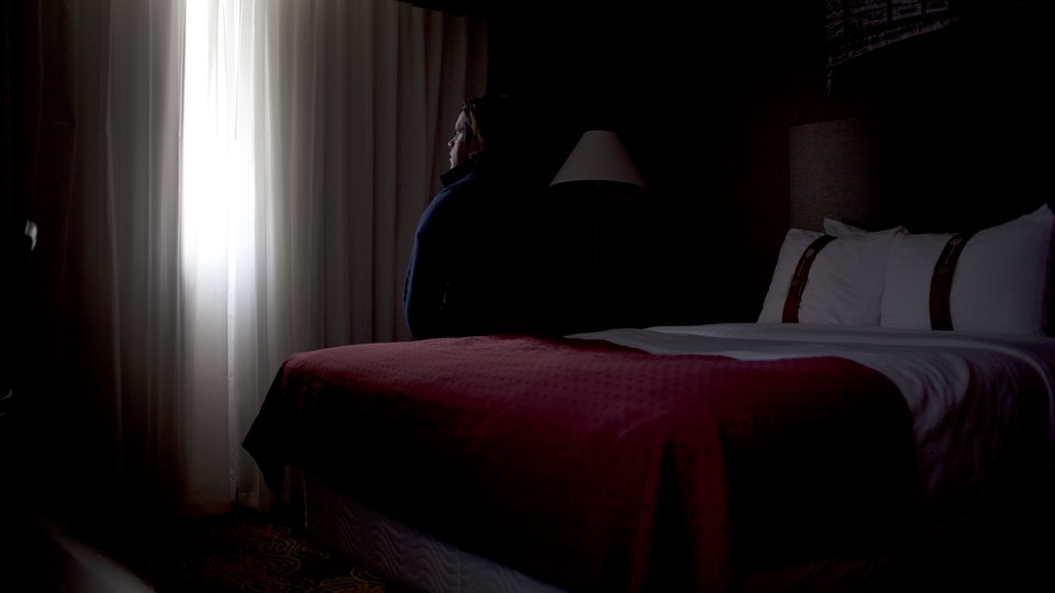 A young man sits on a hotel-room bed, alone, and looks out the window.