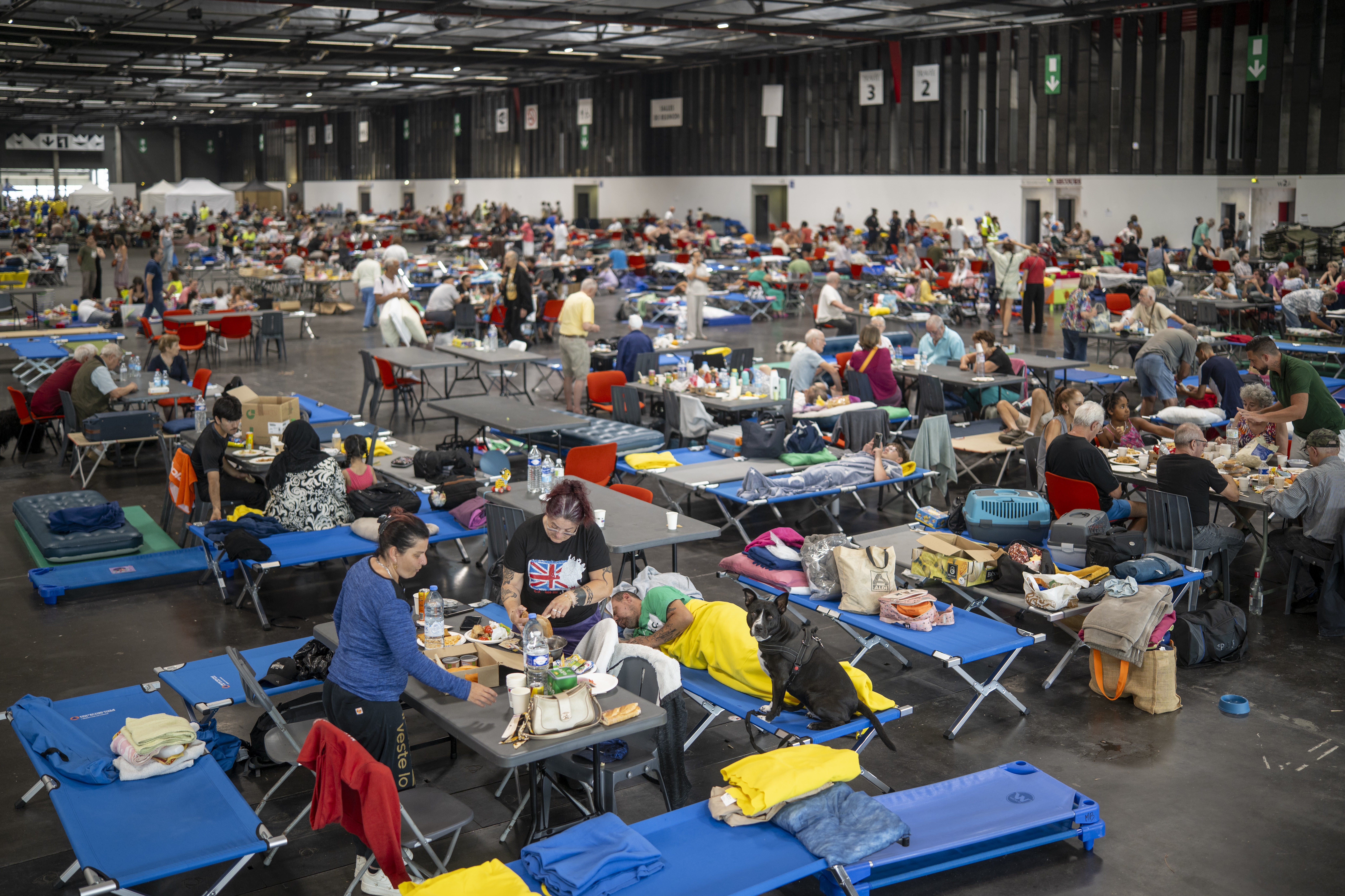 People gather in a temporary shelter building, sitting at tables and on cots.