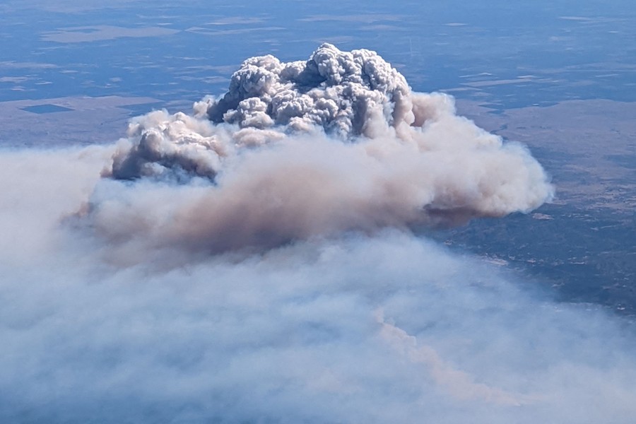 A view from an aircraft showing a large plume of smoke rising above a forest fire.