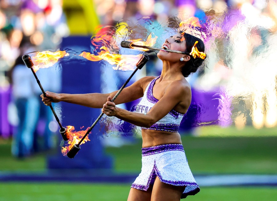 A person performs a routine with three flaming batons, one of which is held in their teeth.