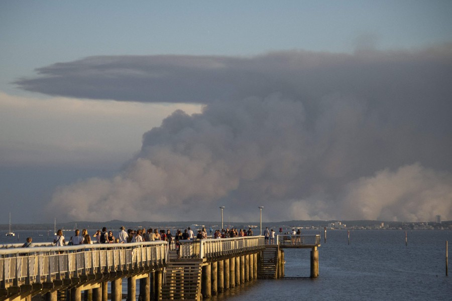People gather on an ocean pier to look toward a huge plume of dark smoke in the distance.