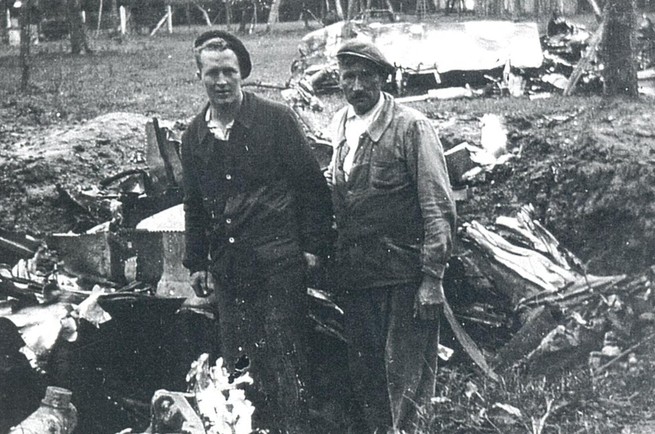 Black and white photo of two men, one young, one old, standing in front of the wreckage of a plane