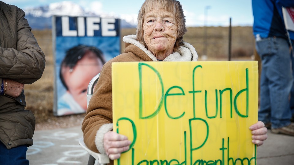 An older woman holds a yellow sign that says "Defund Planned Parenthood" in green letters, with a mountain in the background and people standing beside her