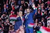 Victor Orbán stands next to J. D. Vance as both of them raise one hand to wave to the crowd.