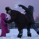 An adult comforts one child while another throws a snowball behind them.