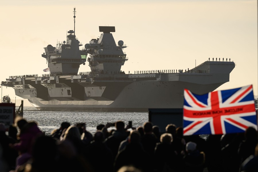A crowd looks out at sailors standing on the deck of a new aircraft carrier just offshore.