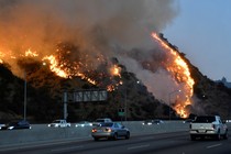 A fire burns on a hillside next to a freeway as cars drive by.