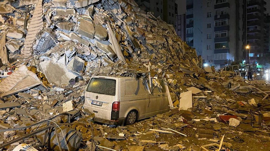 A large pile of rubble sits among buildings and half-covers a van.