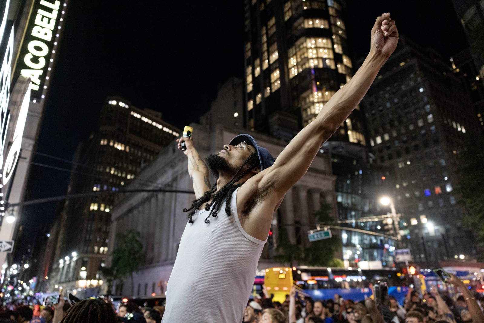 Knicks fans attend a watch party in a city street, with one fan standing at center, arms raised up.