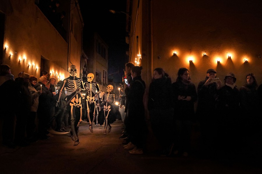 Several people in skeleton costumes walk down a narrow street as people watch.