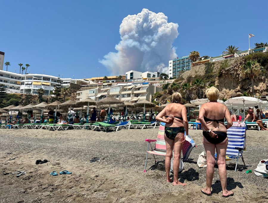 Beachgoers at a resort look inland, toward a towering plume of smoke in the distance.