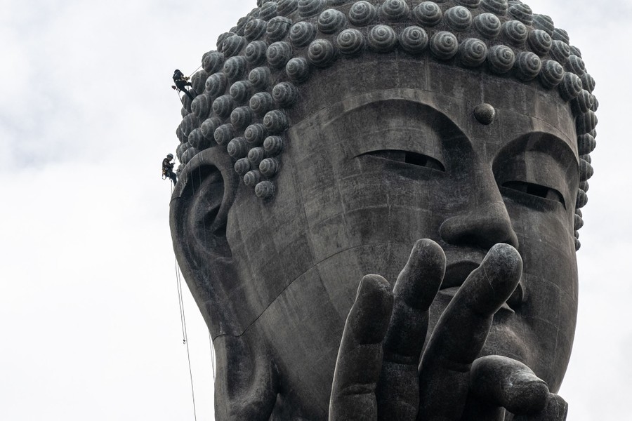 Two workers hang from ropes as they wash part of the head of a large statue of Buddha.