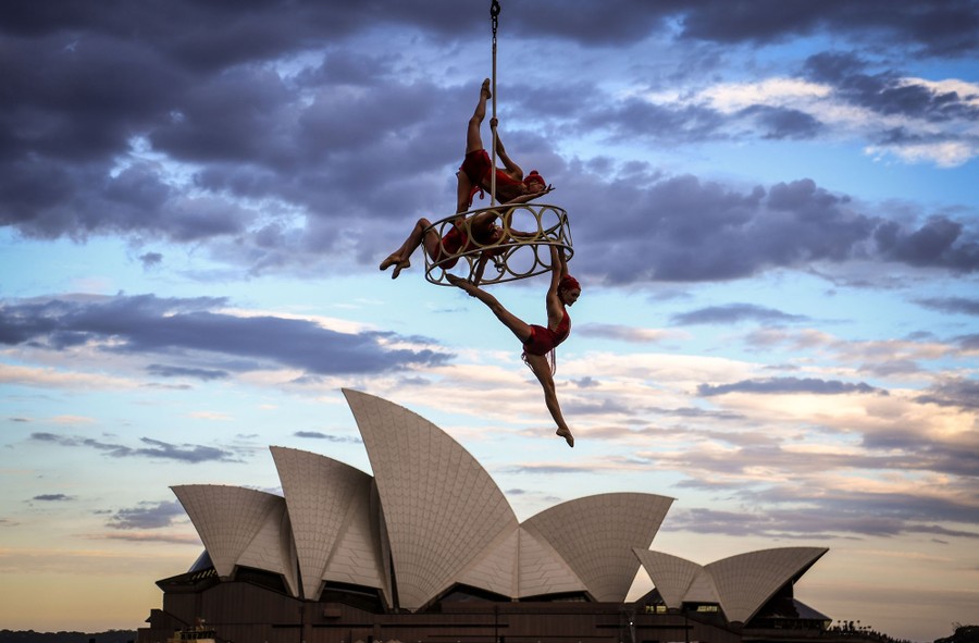 Three performers hang from and on an apparatus suspended by a cable, in front of the Sydney Opera House.