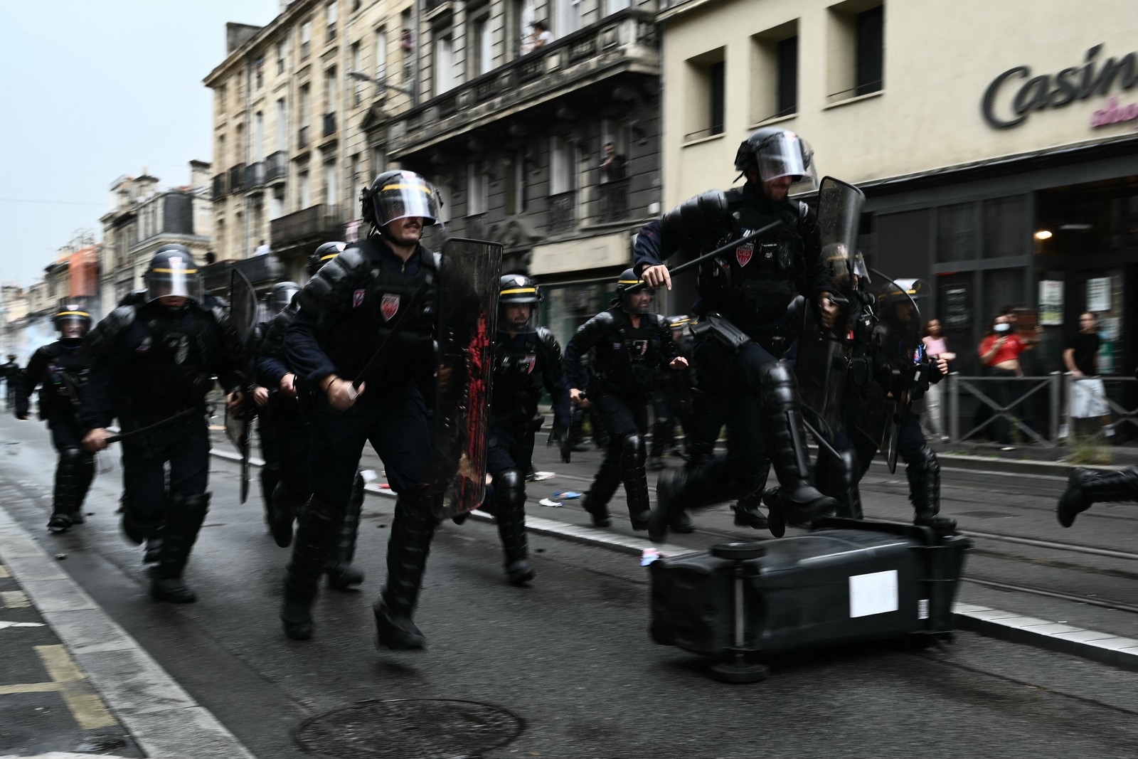 A group of riot police in full gear run down a street. One hurdles a toppled garbage bin.
