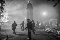 Black-and-white photo of armed troopers in full body armor and gas masks in foggy city park near statue on tall pedestal