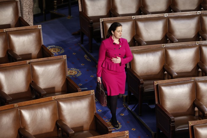 Elise Stefanik wears a pink suit and walks in front of empty seats in the House Chamber