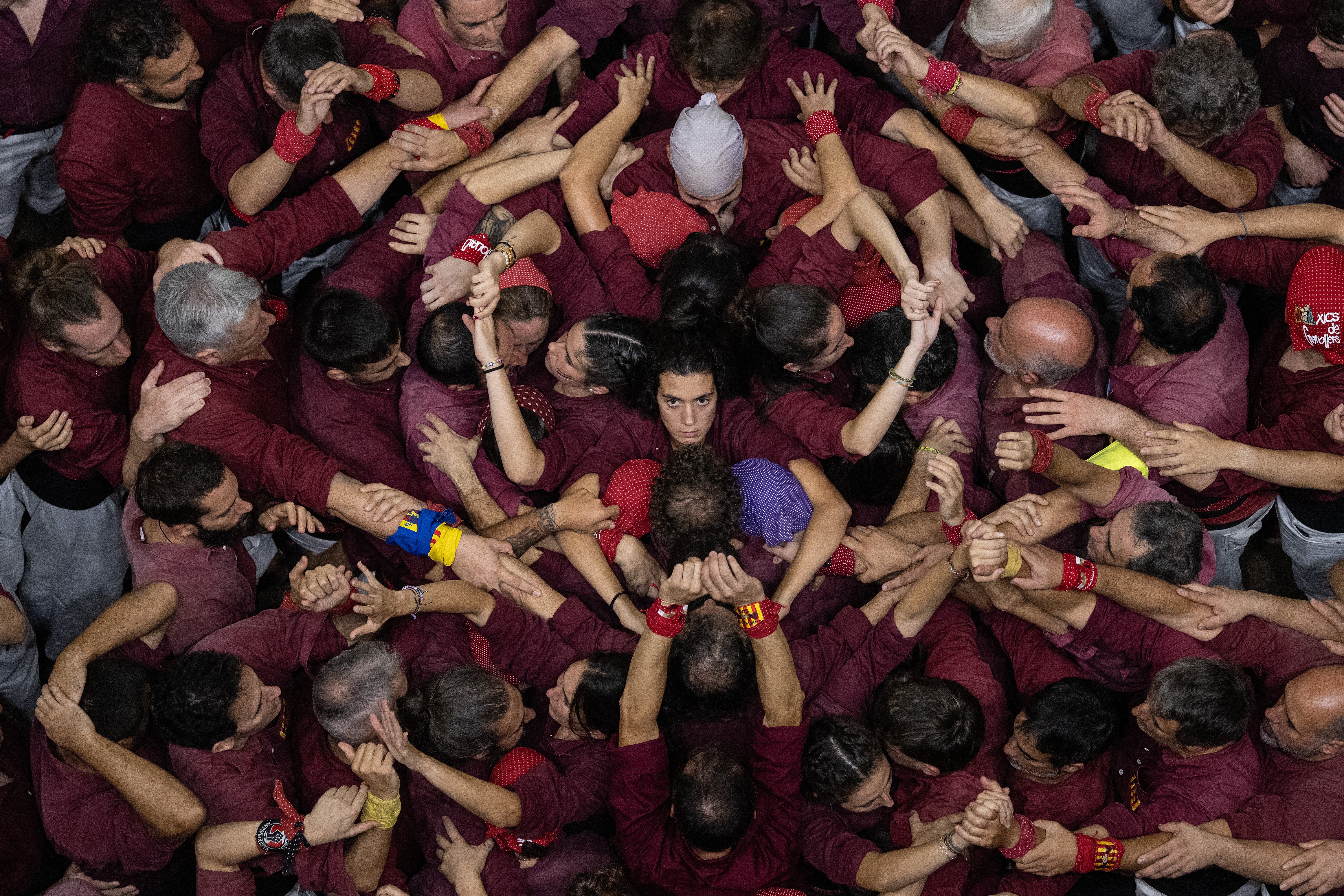 An overhead view of a group of people wearing purple shirts, preparing to build a human tower. One person at center looks straight up toward the camera.