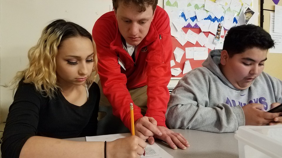 A teacher leans over two students, one of whom is holding a pencil and writing on a worksheet.