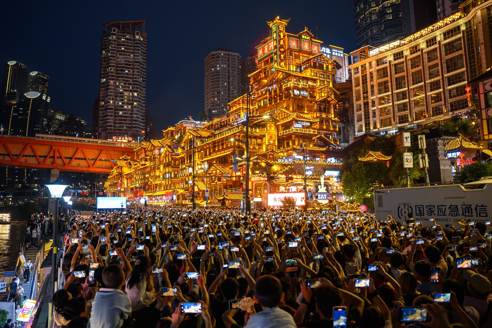 Dozens of people in a crowd hold up their phones to take images of a brightly-lit ornate building in a city.