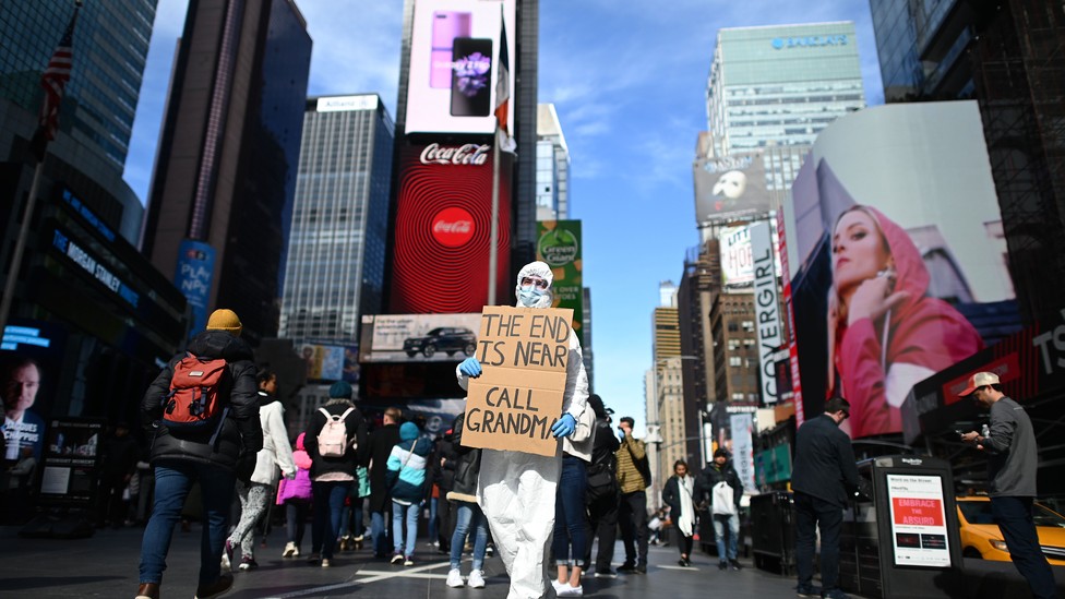 A person in protective gear holds a sign that says "THE END IS NEAR CALL GRANDMA."