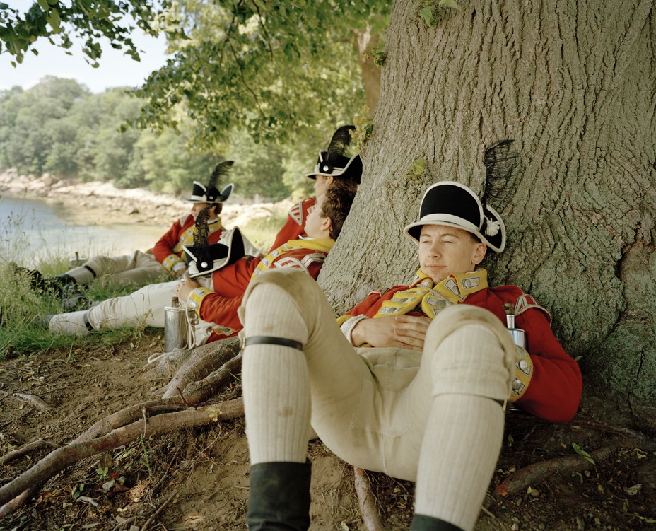 photo of several reenactors dressed as British soldiers sitting in the shade on grass and leaning against the base of a very large tree during a pause, with the shore in background