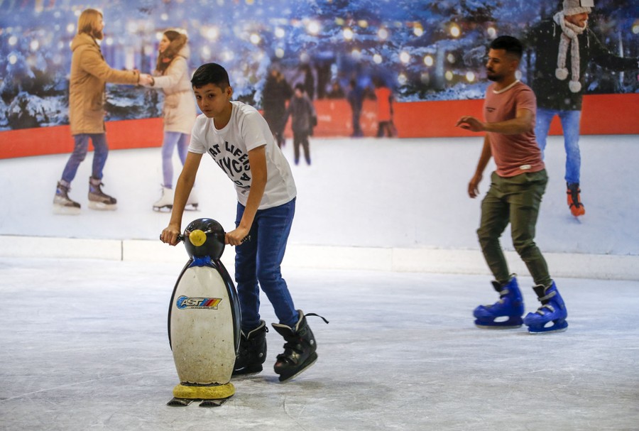 People skate on an indoor ice rink. One pushes a small penguin statue across the ice.