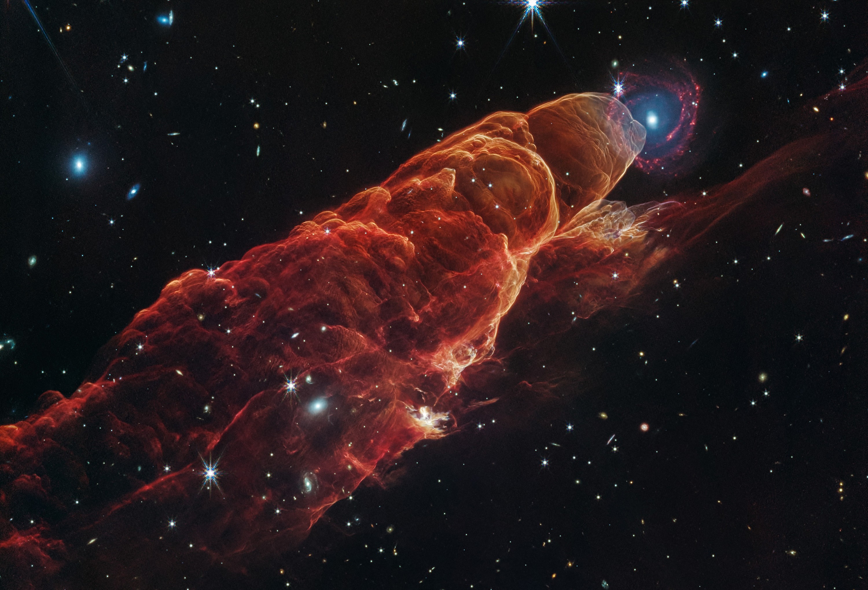 A long, billowing, and glowing cloud of material, seen in deep space, backdropped by stars and more-distant galaxies
