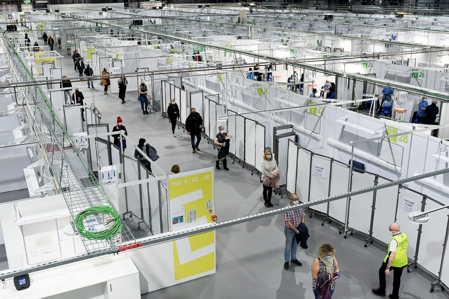 Dozens of people line up, spaced apart, inside a large space filled with temporary vaccination stations.