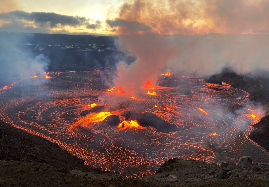 Lava flows within a broad volcanic crater, as steam and gas rise above it.