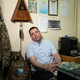 A man wearing a light-blue shirt sits in a chair against the wall in an office.